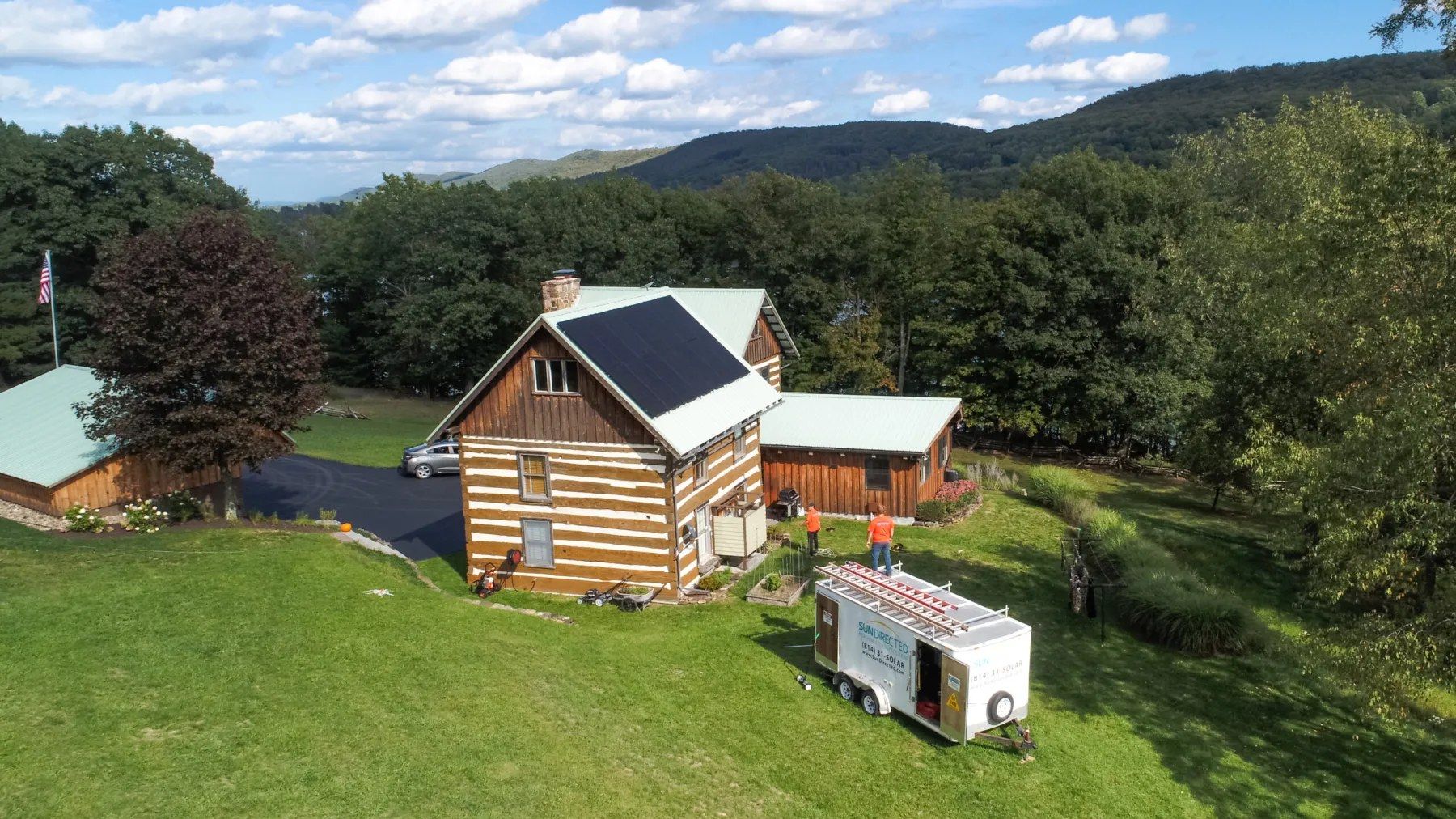 Aerial of a log cabin with rooftop solar and a Sun Directed work trailer on the lawn.